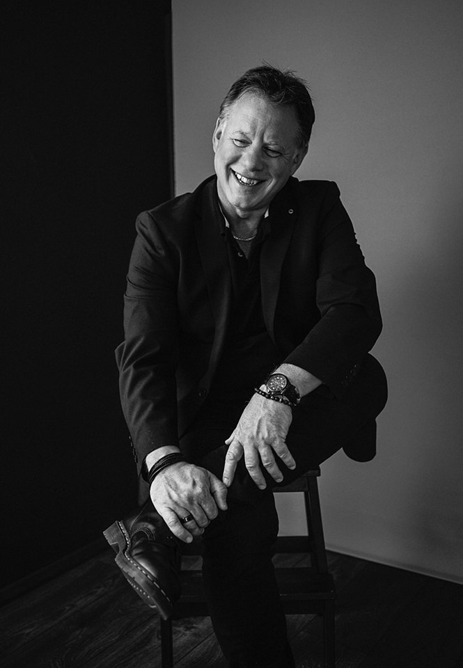 Black and white photo of Paul Ward laughing, seated on a stool in a dark jacket
