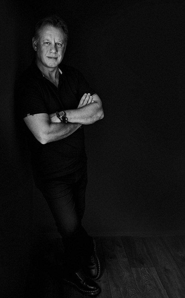 Black and white portrait of Paul Ward standing with his arms folded against a dark wall